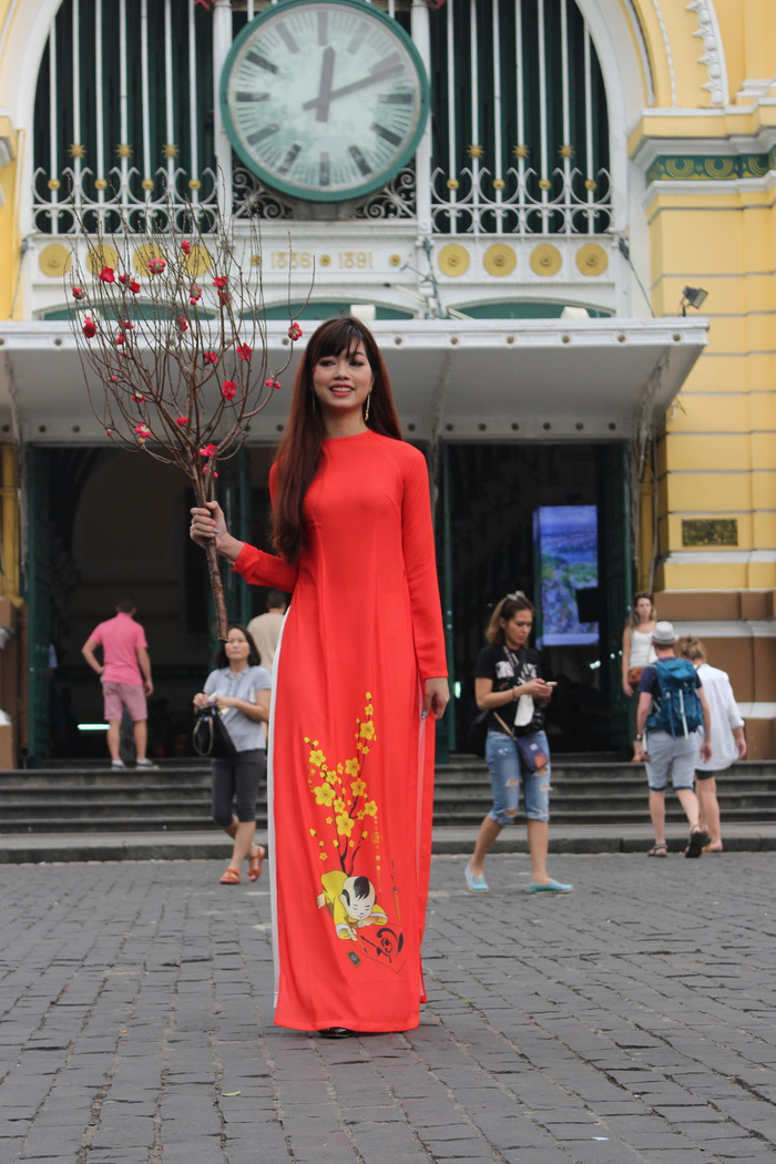 A woman in the traditional ao dai holds a peach branch with the Saigon Central Post Office in the background. Photo: Tuoi Tre