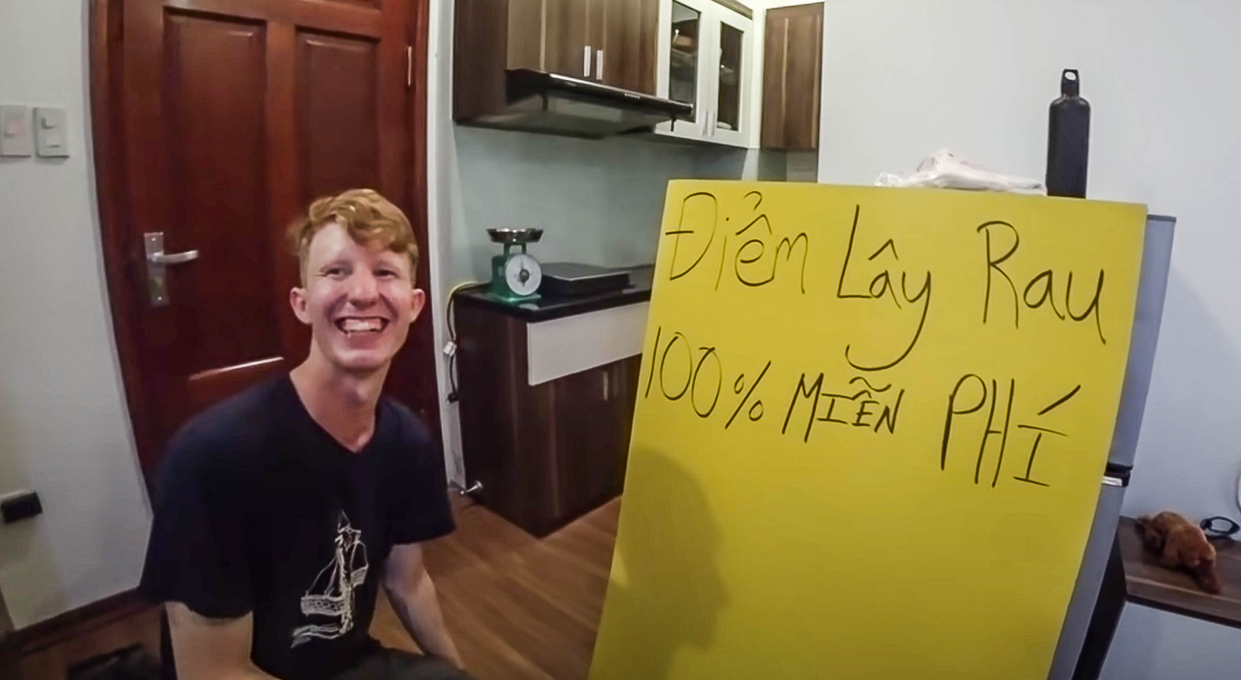 Chris Lewis is seen in a snapshot from his YouTube video, with a board next to him saying '100 percent free vegetable pick up point.' Chris Lewis wrote the sign in Vietnamese himself.