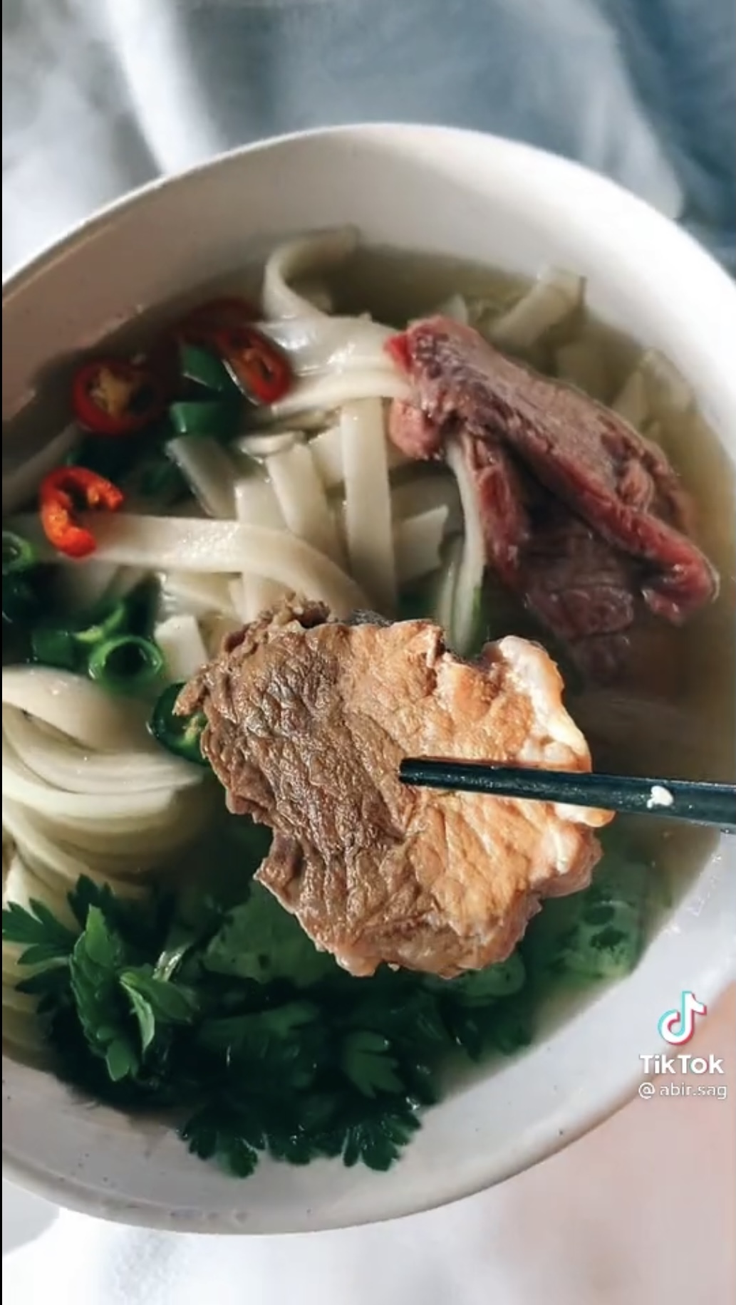 A screenshot shows Abir Saghir putting a piece of beef flank into a bowl of Vietnamese pho she makes in a video on her @abir.sag TikTok channel.