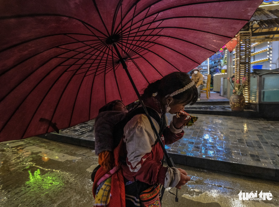 In this image, a girl street vendor, with an umbrella in her hand, is eating a small cake while on way to look for customers in the cold rain on Xuan Vien Street in Sa Pa Town.