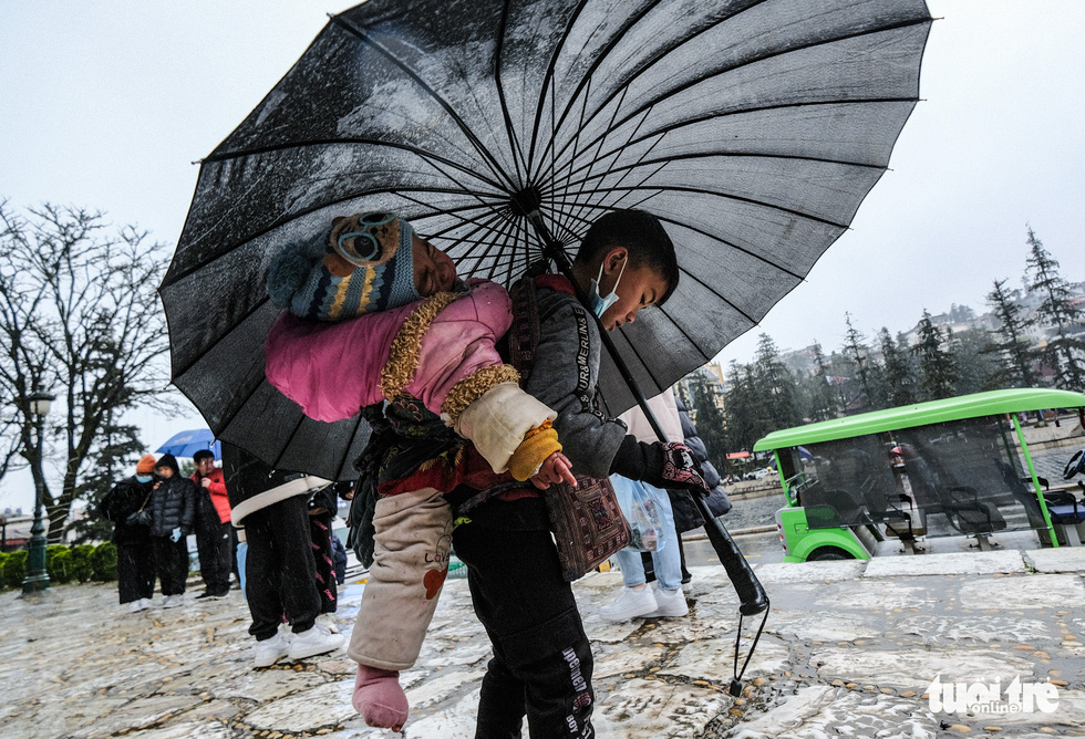 A child peddler, carrying a crying little girl on his back, is seen going with an umbrella in the rain at the Stone Church in Sa Pa Town in this image.