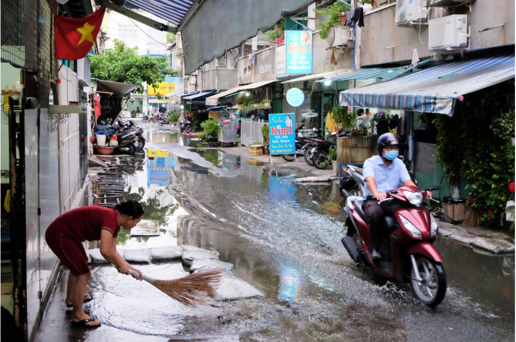 A motorbike wades through a flooded route while a woman tries to sweep stagnant water away at An Quang apartment building. Photo: Phuong Nhi / Tuoi Tre